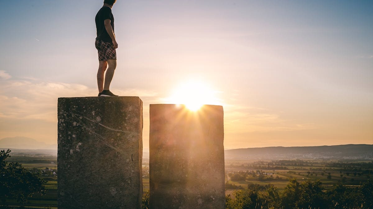 A man stands on stone blocks at sunset, gazing over a scenic Austrian landscape, evoking themes of solitude and exploration.
